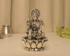 Large sterling silver Lakshmi idol on a broad lotus base, on a pale table with a silver pot behind.