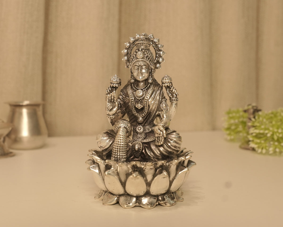 Large sterling silver Lakshmi idol on a broad lotus base, on a pale table with a silver pot behind.