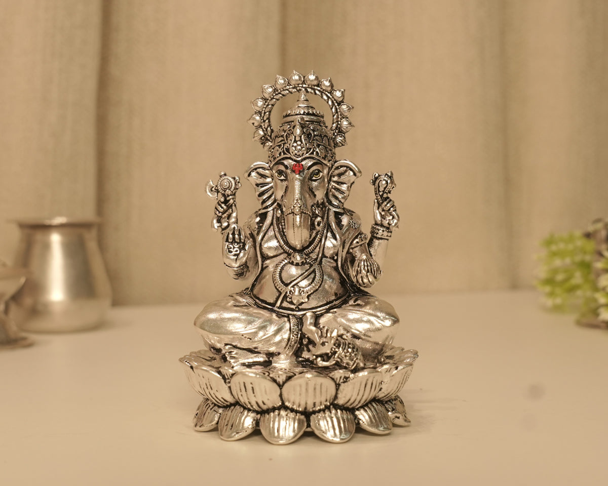 Large sterling silver Ganesh idol on a lotus base, on a pale table with a silver pot behind.