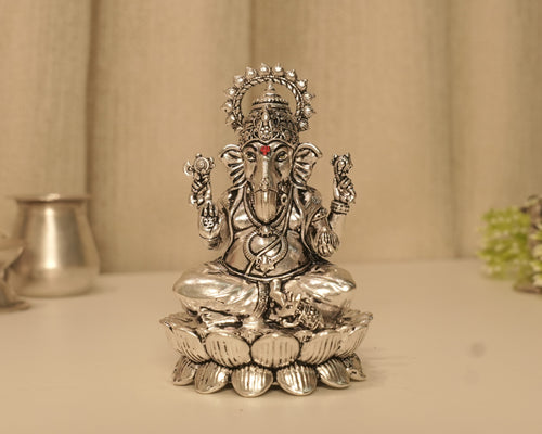 Large sterling silver Ganesh idol on a lotus base, on a pale table with a silver pot behind.