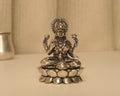 Sterling silver Lakshmi idol on a lotus base, on a pale table with a silver pot behind.