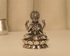 Sterling silver Lakshmi idol on a lotus base, on a pale table with a silver pot behind.