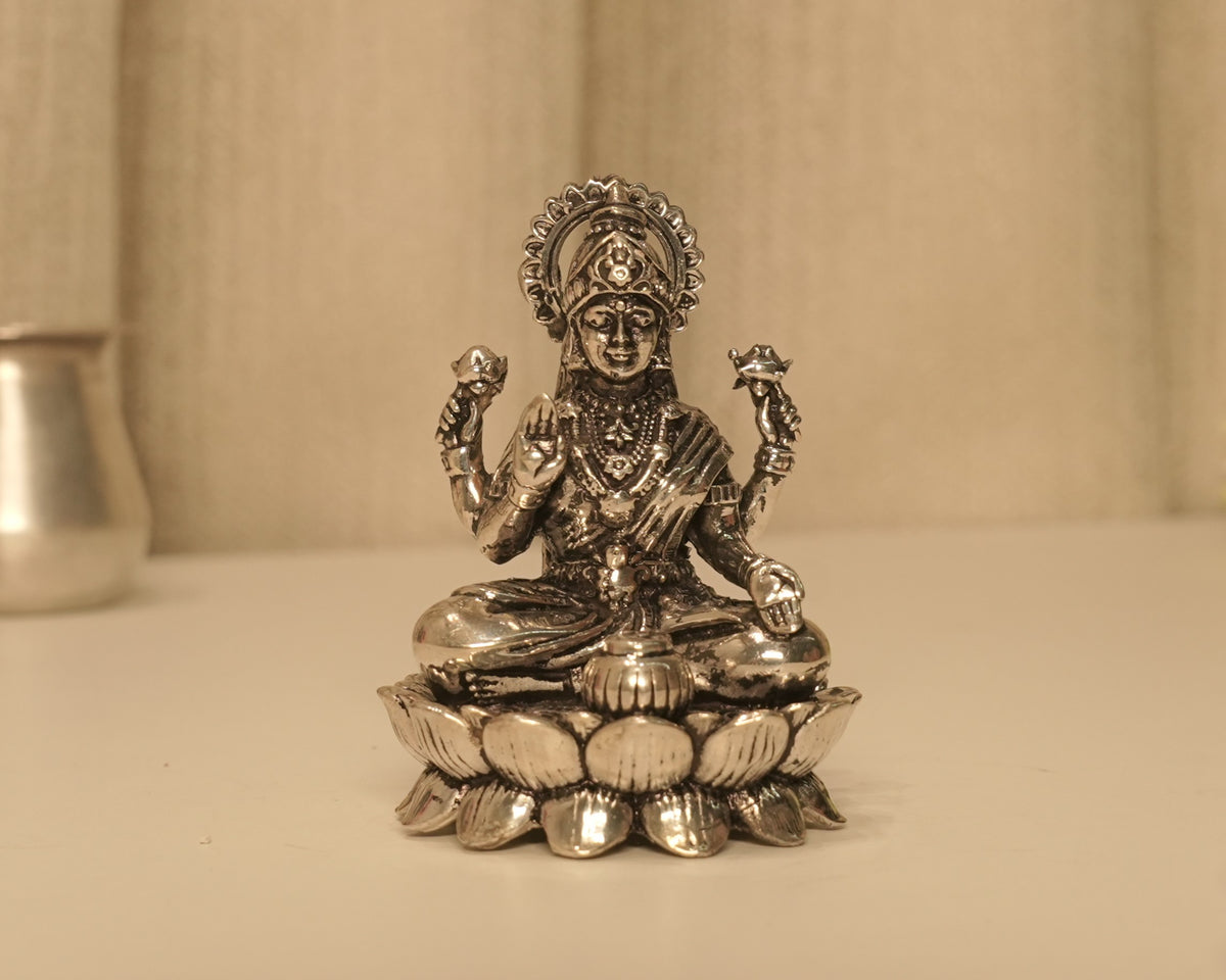 Sterling silver Lakshmi idol on a lotus base, on a pale table with a silver pot behind.