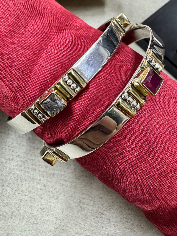 Sterling silver bangles resting on red silk, one station holding a deep red square stone and one a colourless stone in gold-tone bezels.