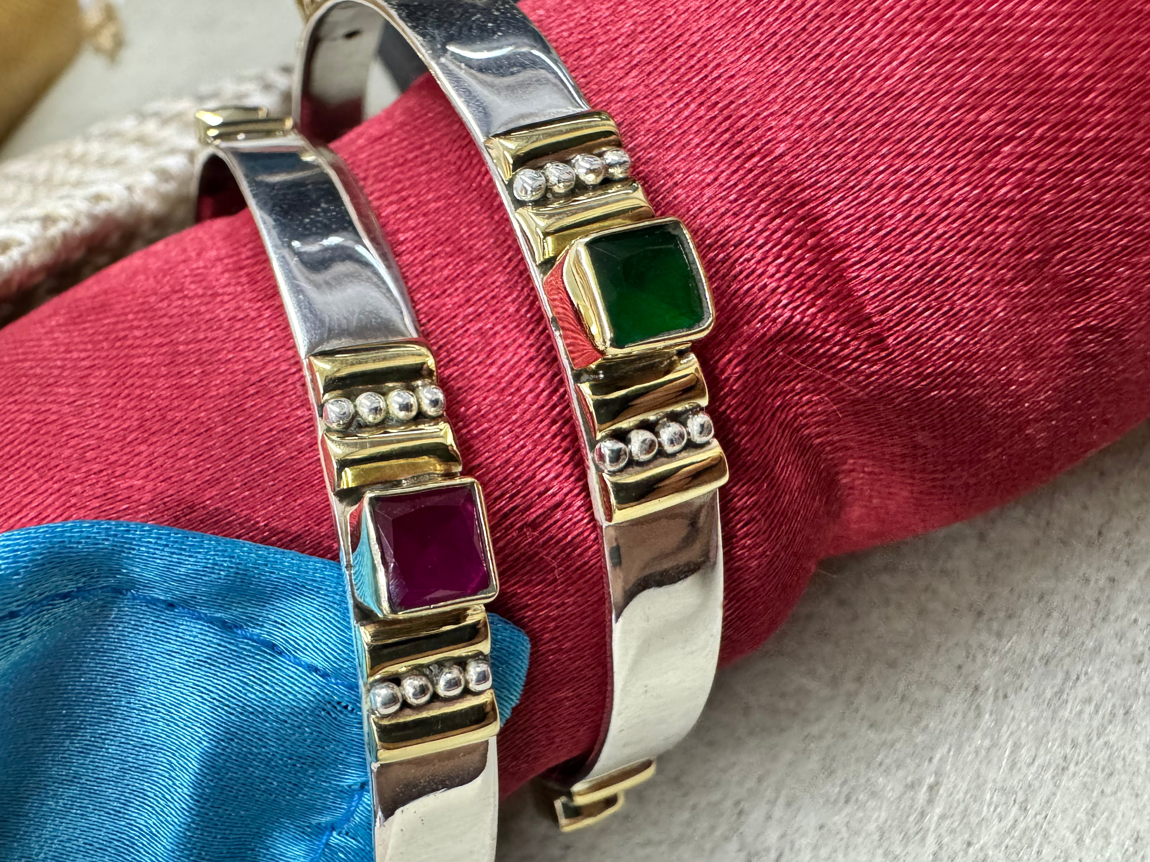 Close view of green and deep red square stones in gold-tone bezels on the polished sterling silver bangles, against red silk.