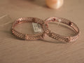 Pair of rose-tone bangles with criss cross lattice panels and American Diamond rails, lying together on a shelf.