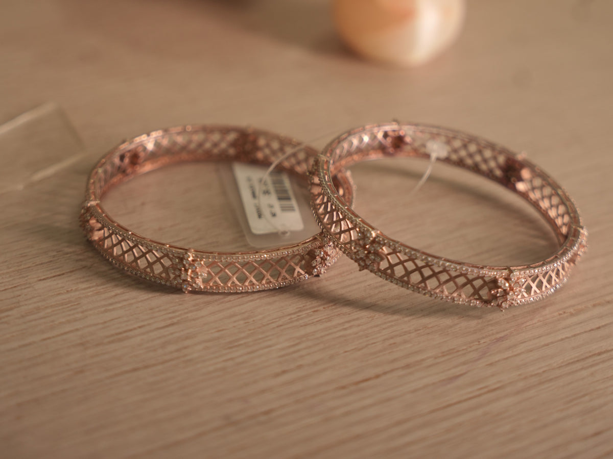 Pair of rose-tone bangles with criss cross lattice panels and American Diamond rails, lying together on a shelf.