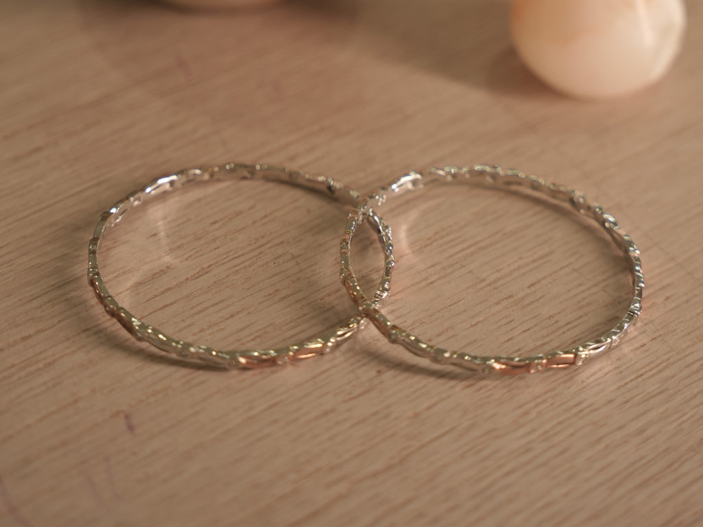 Pair of slim sterling silver bangles lying flat, showing the ribbon links and rose-tone touches around the band.