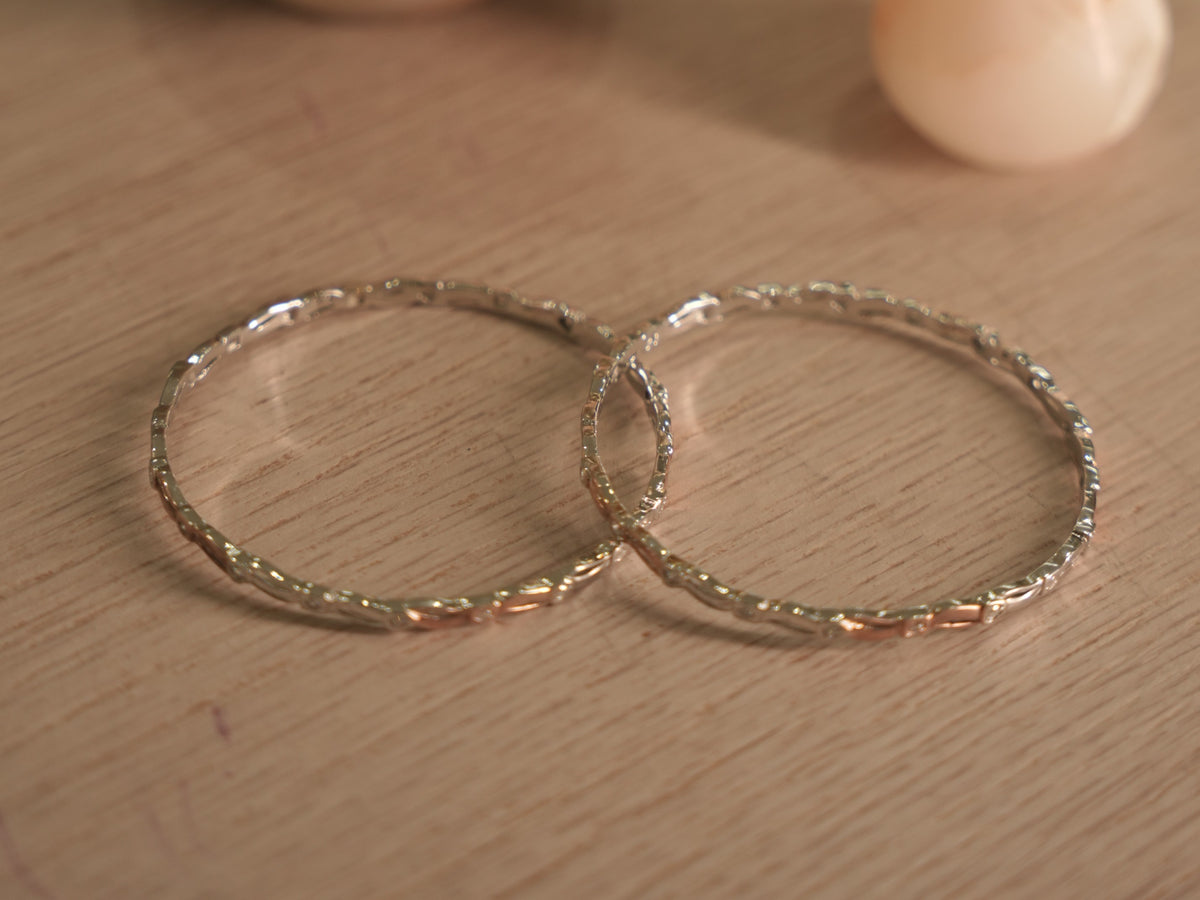 Pair of slim sterling silver bangles lying flat, showing the ribbon links and rose-tone touches around the band.