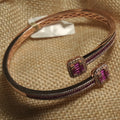 Rose gold-tone bypass bangle with a channel of magenta baguette cubic zirconia bordered in white Bejewelled, ending in two halo squares, resting on a jute pouch.