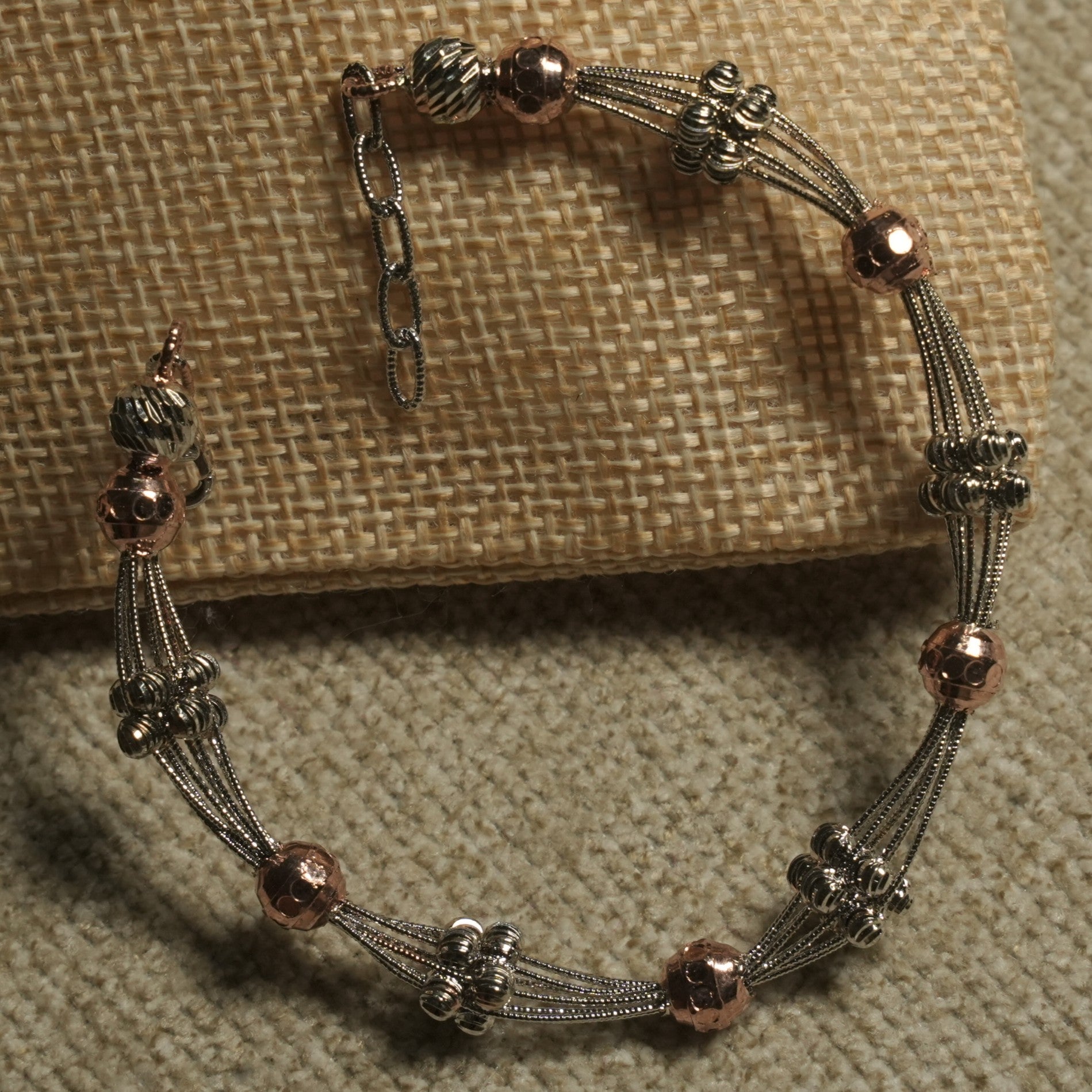 Multi-strand textured silver bracelet stationed with fluted silver beads and hammered rose gold-tone beads, with extender chain, resting on a jute pouch.