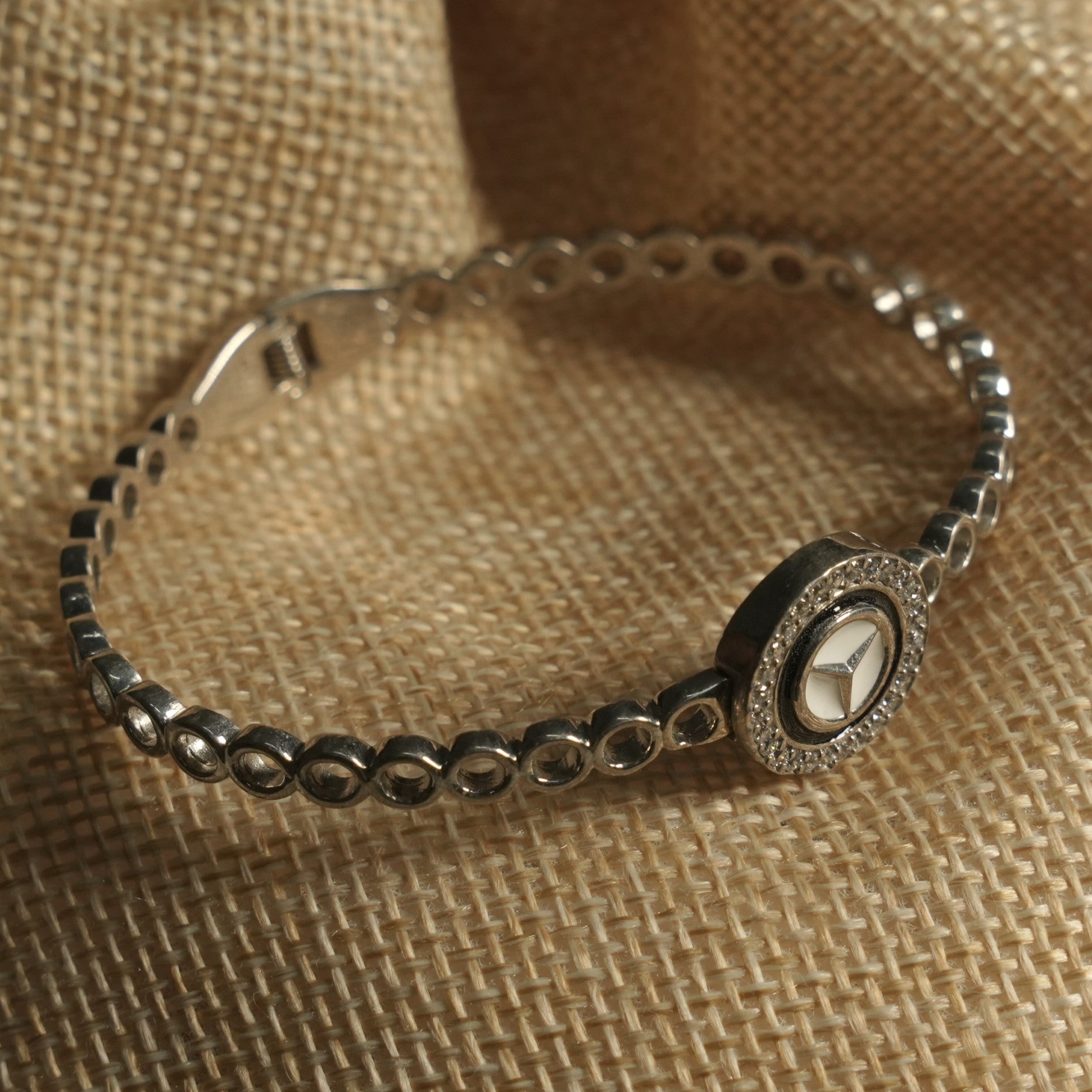 Sterling silver bracelet with an open bubble-link band and a round medallion of white enamel with a star motif ringed by Bejewelled cubic zirconia, on a jute pouch.
