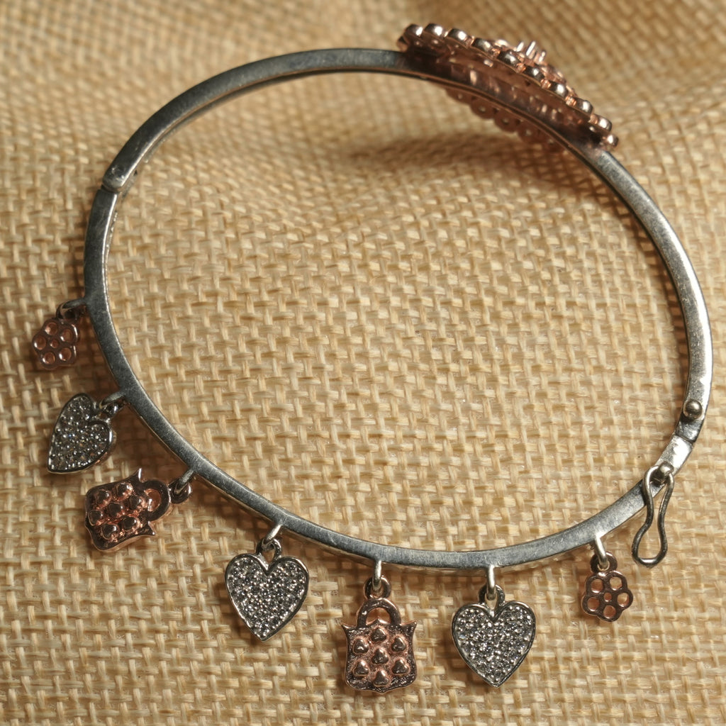 Silver-tone hinged bangle hung with Bejewelled heart, blossom and rose gold-tone charms, resting on a jute pouch.