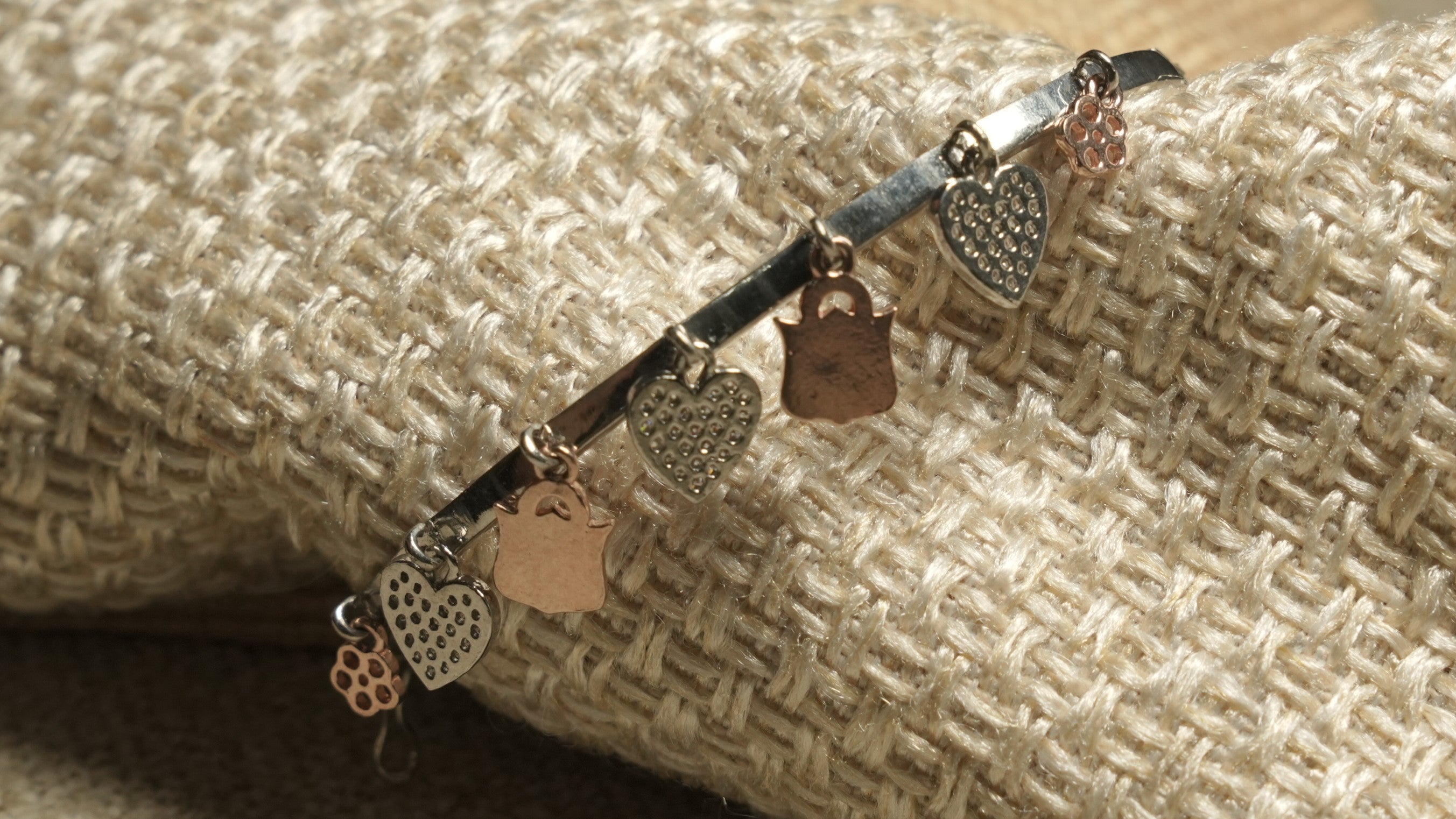 Detail of the bangle's dangling charms, including Bejewelled hearts and rose gold-tone drops, against woven fabric.