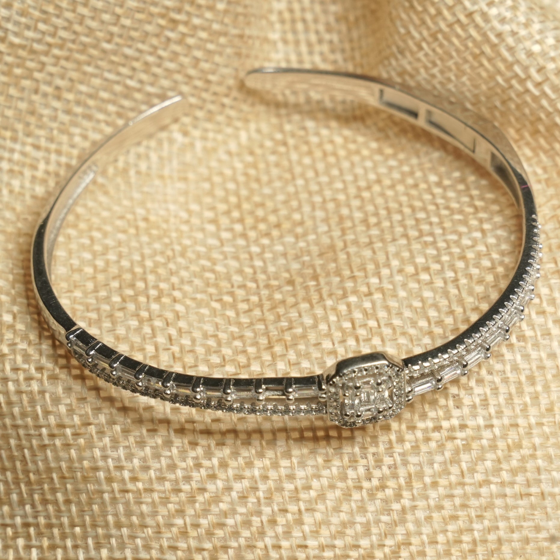 Sterling silver hinged bangle with a channel of baguette cubic zirconia, micro-Bejewelled borders and a square halo centre, resting on a jute pouch.
