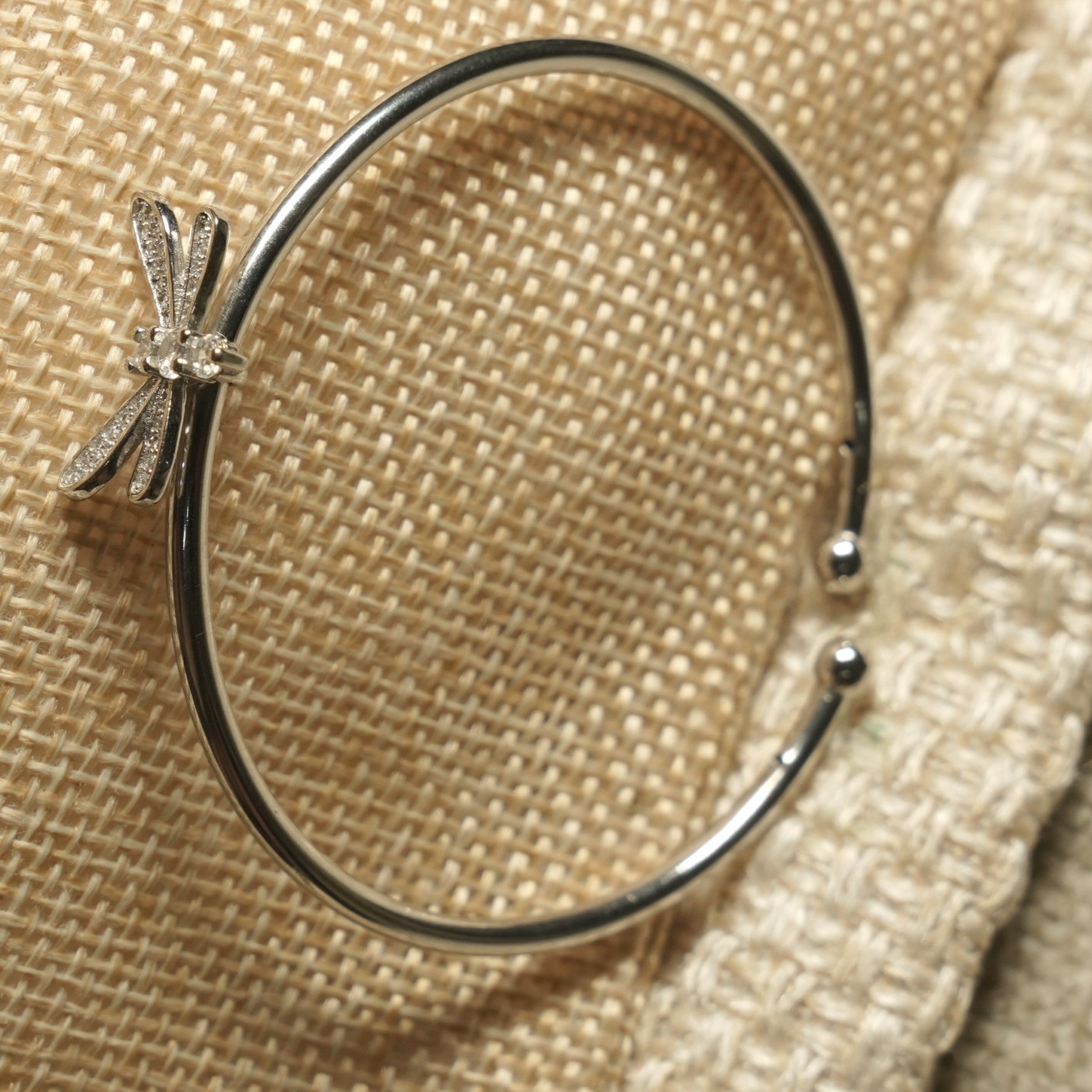 Polished sterling silver open cuff with ball ends and a cubic zirconia Bejewelled dragonfly perched on one side, on woven jute fabric.
