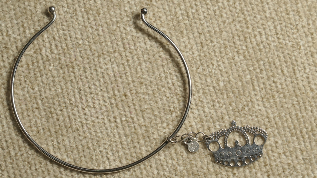 Slim sterling silver wire cuff with ball ends and a dangling openwork crown charm, laid on beige fabric.