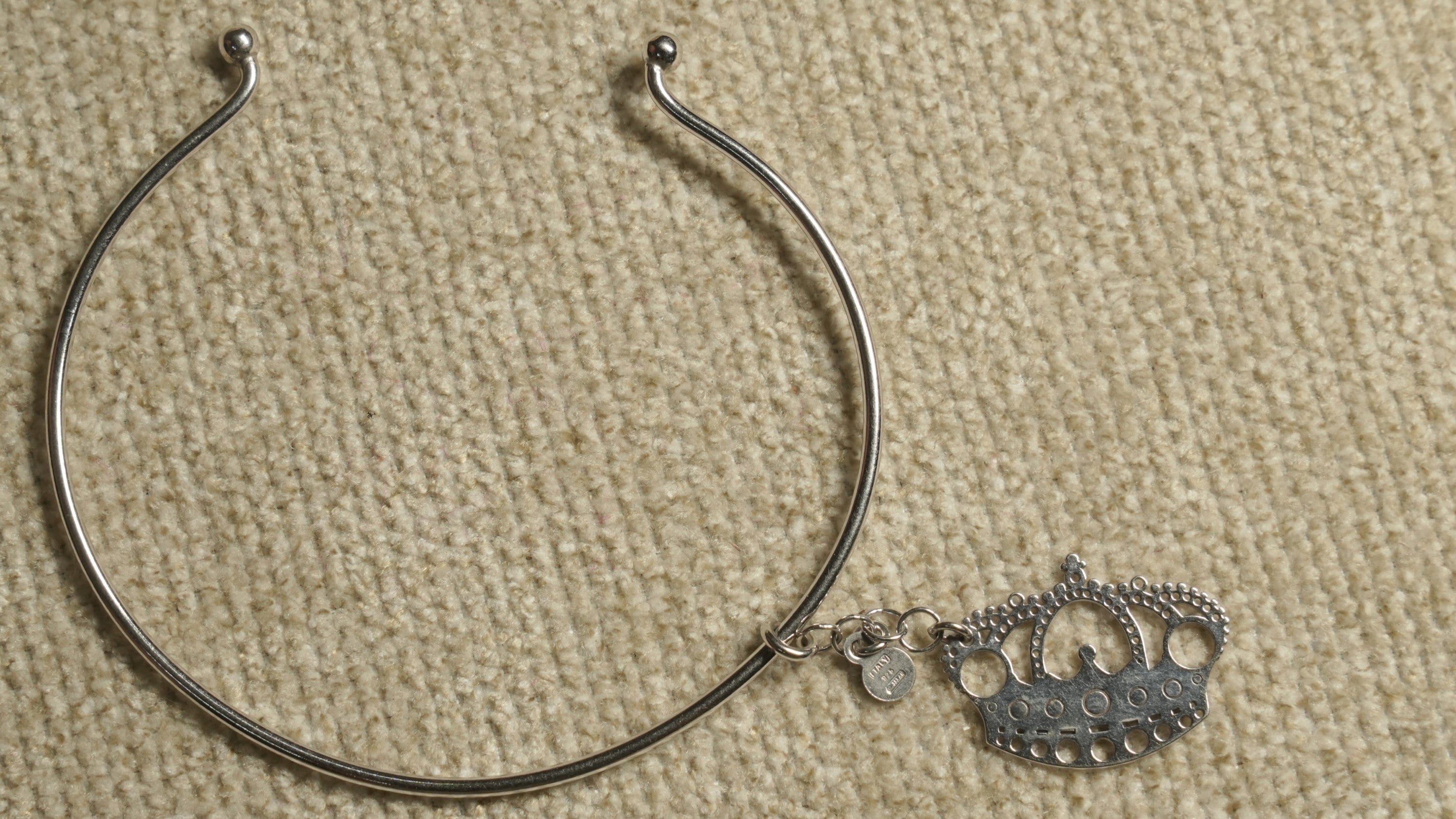 Slim sterling silver wire cuff with ball ends and a dangling openwork crown charm, laid on beige fabric.