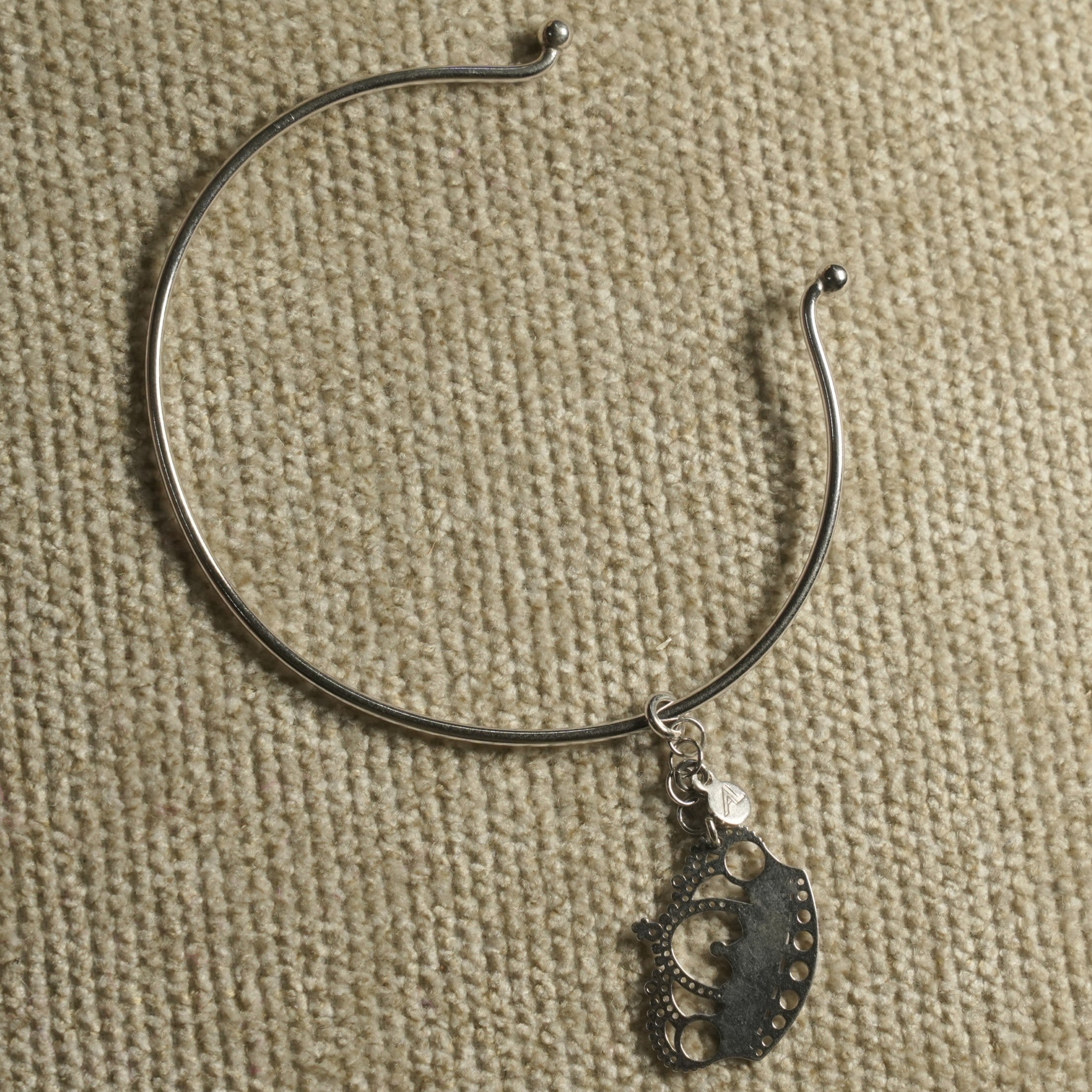 The wire cuff turned on woven fabric, showing the pierced crown charm and small round tag on its link.