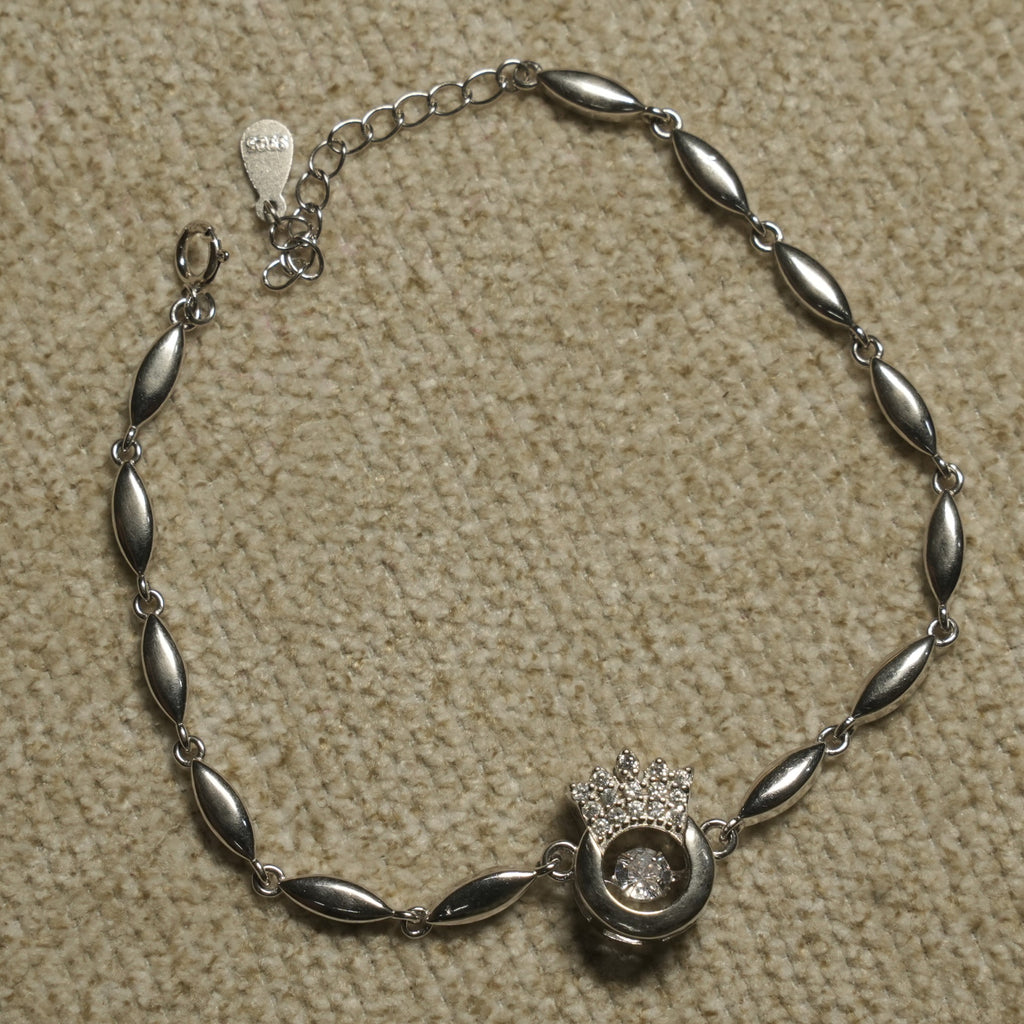 Sterling silver bracelet of marquise-shaped links centred on an open circle with a Bejewelled crown and a suspended cubic zirconia, laid flat on beige fabric.