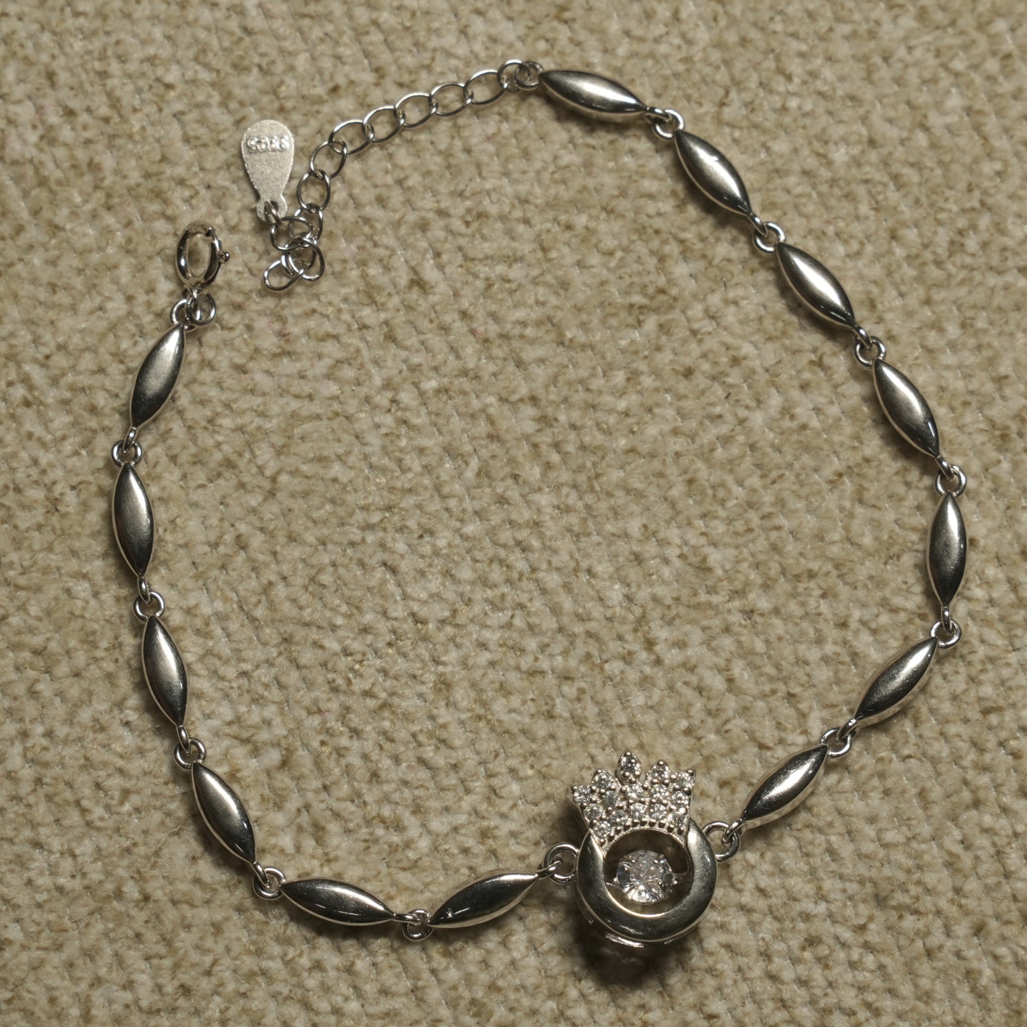 Sterling silver bracelet of marquise-shaped links centred on an open circle with a Bejewelled crown and a suspended cubic zirconia, laid flat on beige fabric.