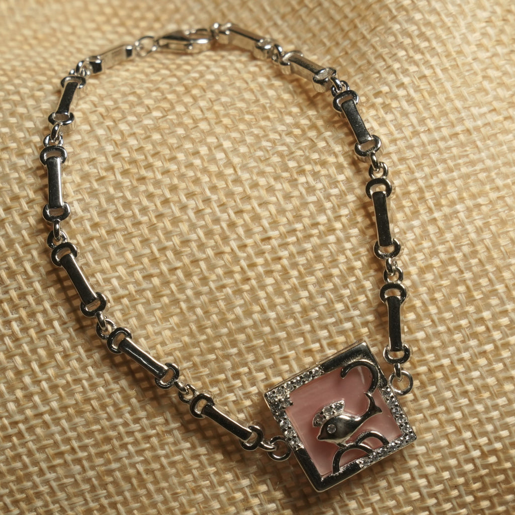 Sterling silver bar-link bracelet with a square pearlescent pink panel framed in cubic zirconia and centred with a fish motif, resting on a jute pouch.