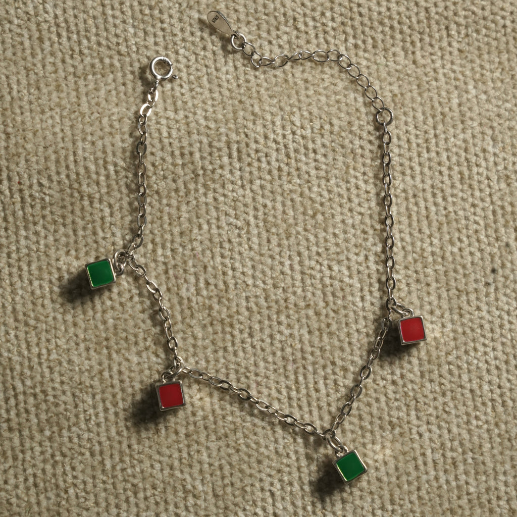Fine sterling silver cable chain bracelet with alternating red and green enamel square charms and a spring-ring clasp, on beige fabric.