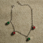 Fine sterling silver cable chain bracelet with alternating red and green enamel square charms and a spring-ring clasp, on beige fabric.