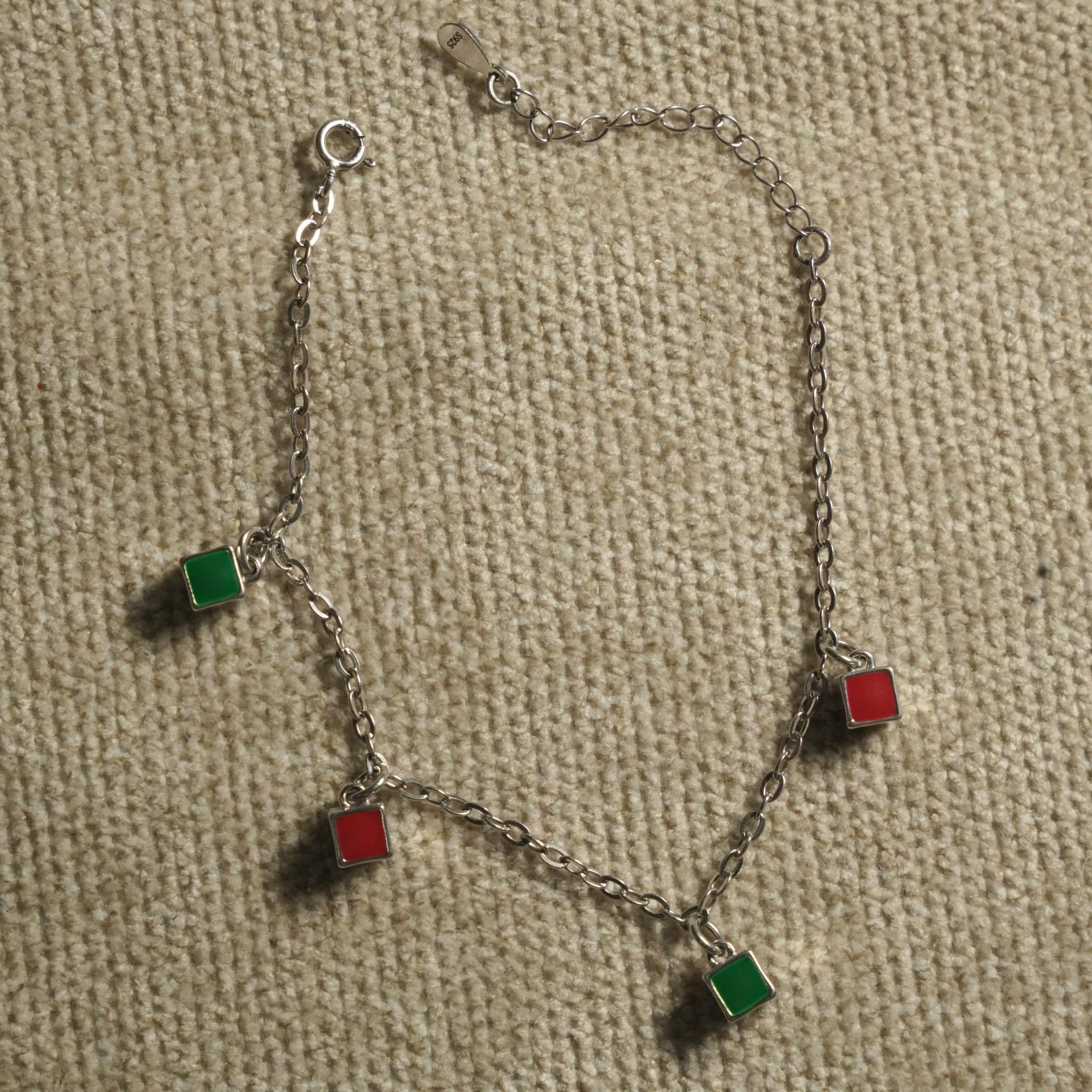 Fine sterling silver cable chain bracelet with alternating red and green enamel square charms and a spring-ring clasp, on beige fabric.