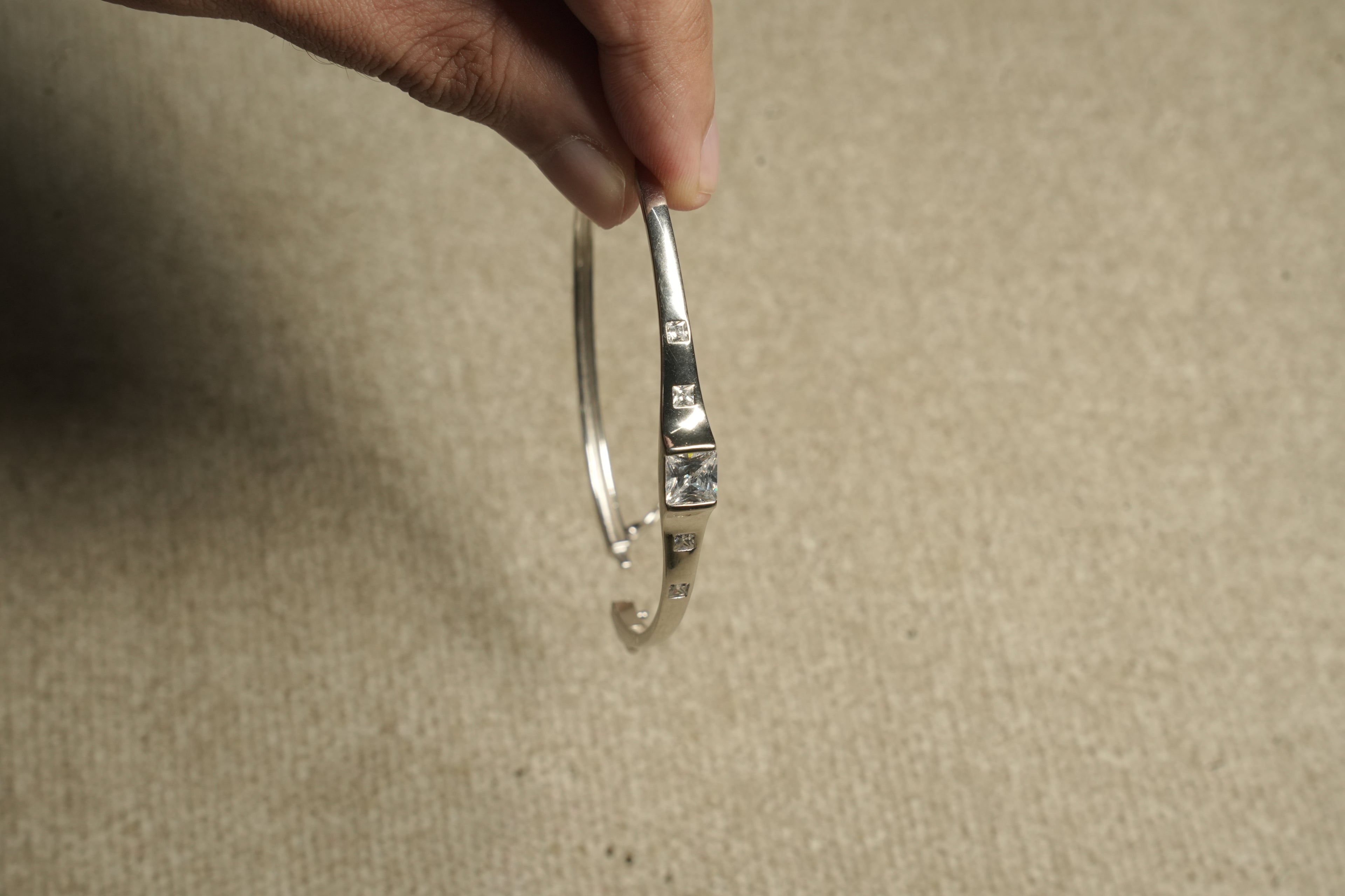 Fine sterling silver kada style bracelet with equal distant square cut stones moving towards the large central squre stone on beige fabric, held in hand