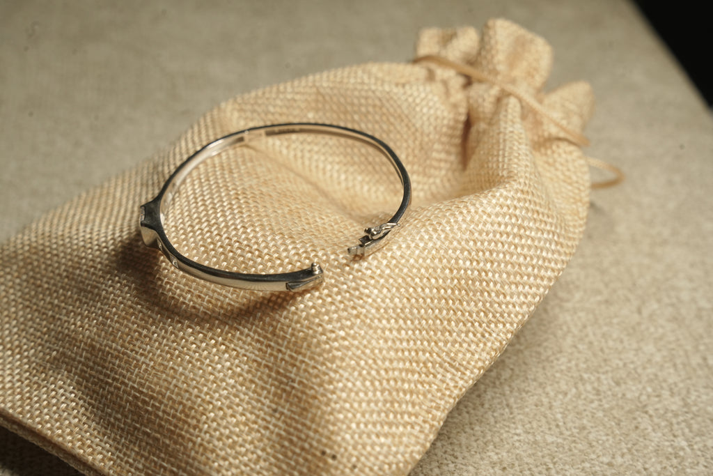 Fine sterling silver kada style bracelet with equal distant square cut stones moving towards the large central squre stone on beige fabric, show lying on a jute bag