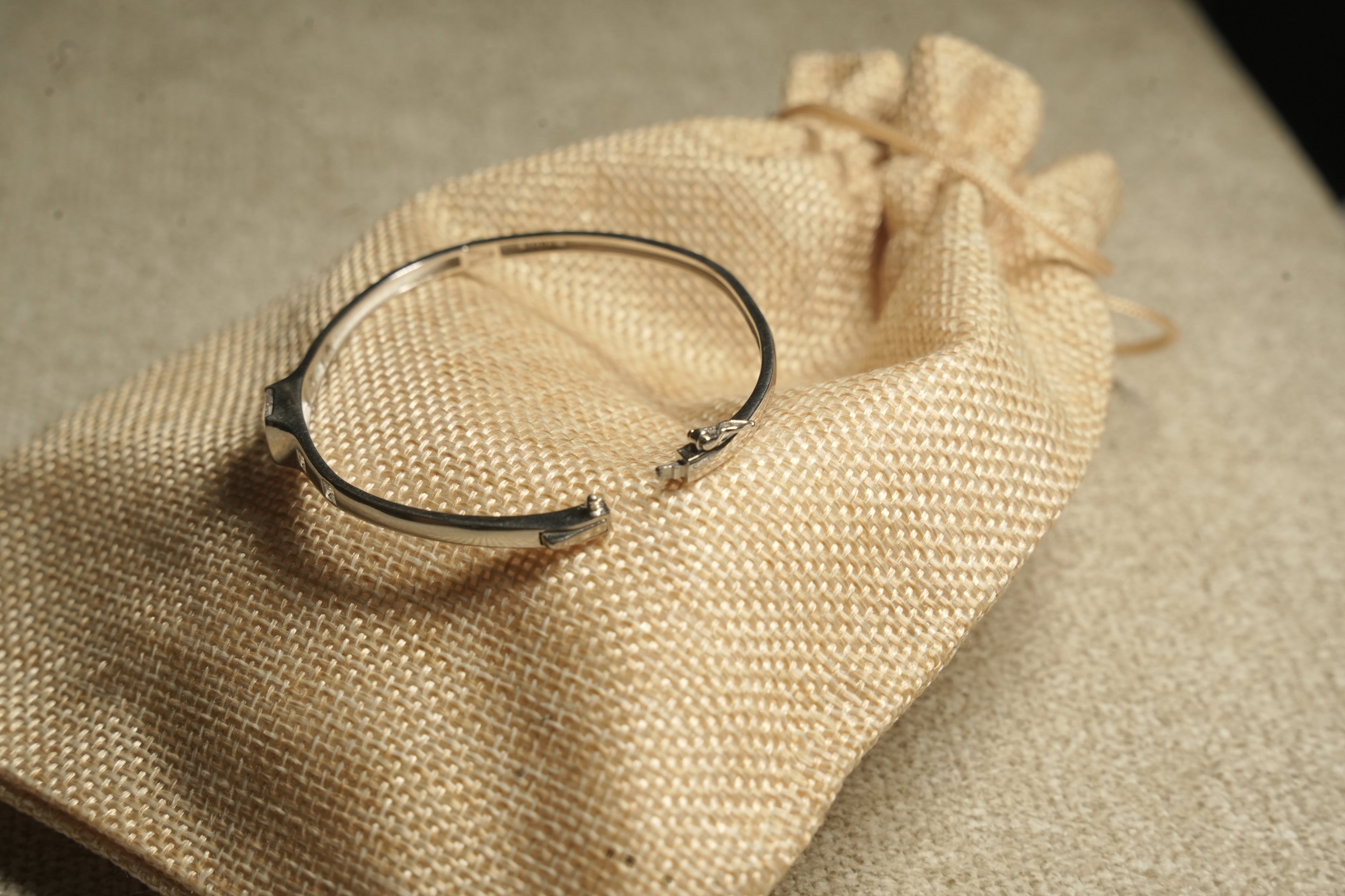 Fine sterling silver kada style bracelet with equal distant square cut stones moving towards the large central squre stone on beige fabric, show lying on a jute bag