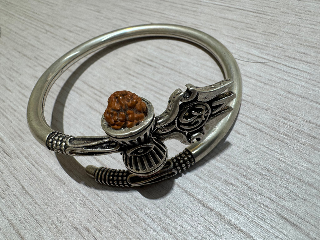 Oxidised sterling silver kada with a trishul, damroo and rudraksha bead, lying on a pale wood surface.