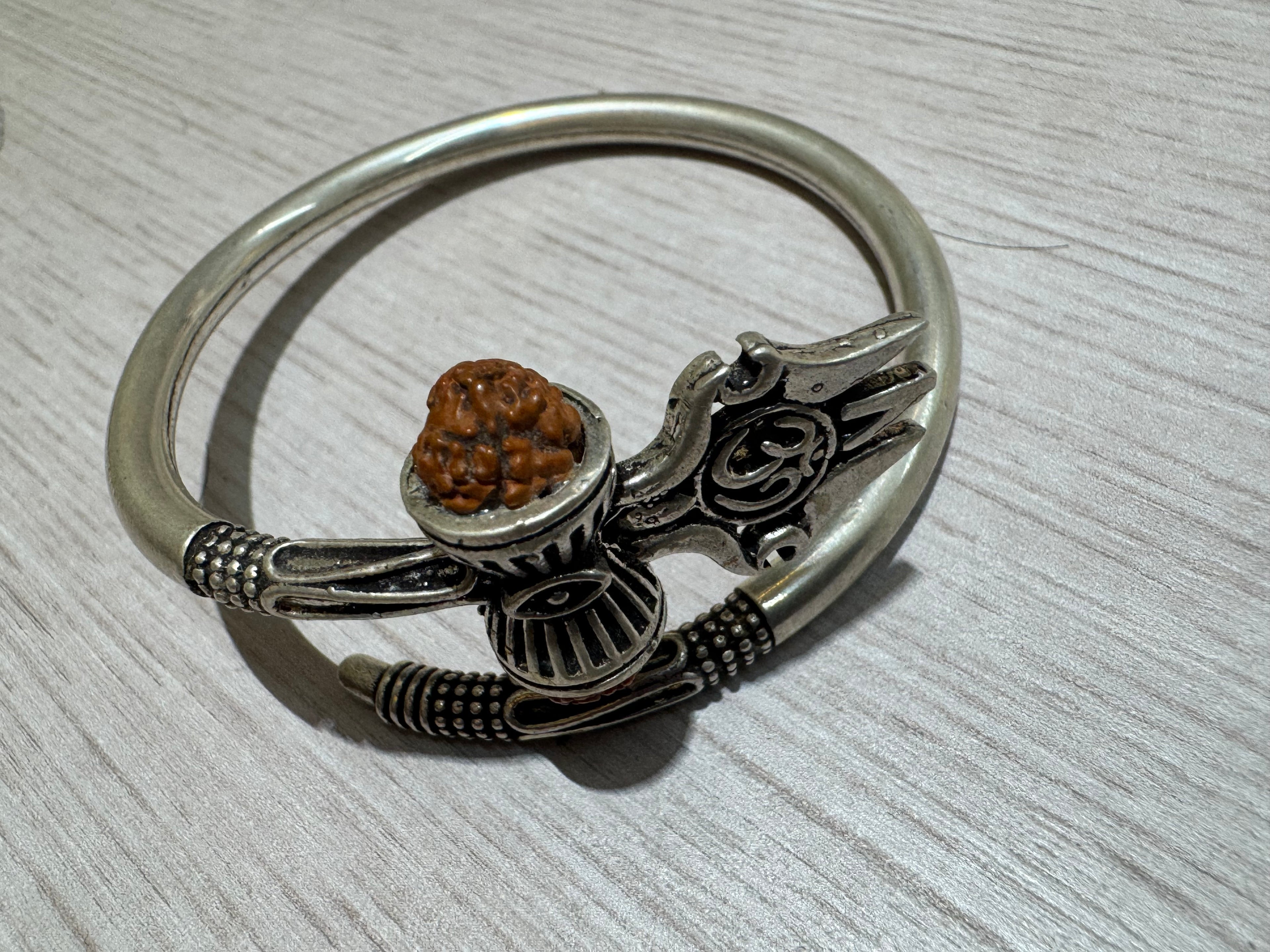 Oxidised sterling silver kada with a trishul, damroo and rudraksha bead, lying on a pale wood surface.