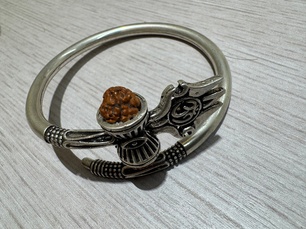 Oxidised sterling silver kada with a trishul, damroo and rudraksha bead, lying on a pale wood surface.