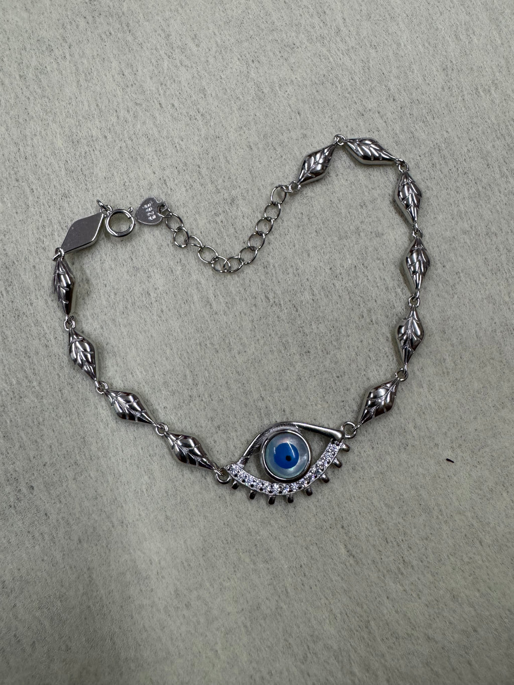 Sterling silver evil eye bracelet arranged on a pale grey ground, the eye at one end and the extender chain at the other.