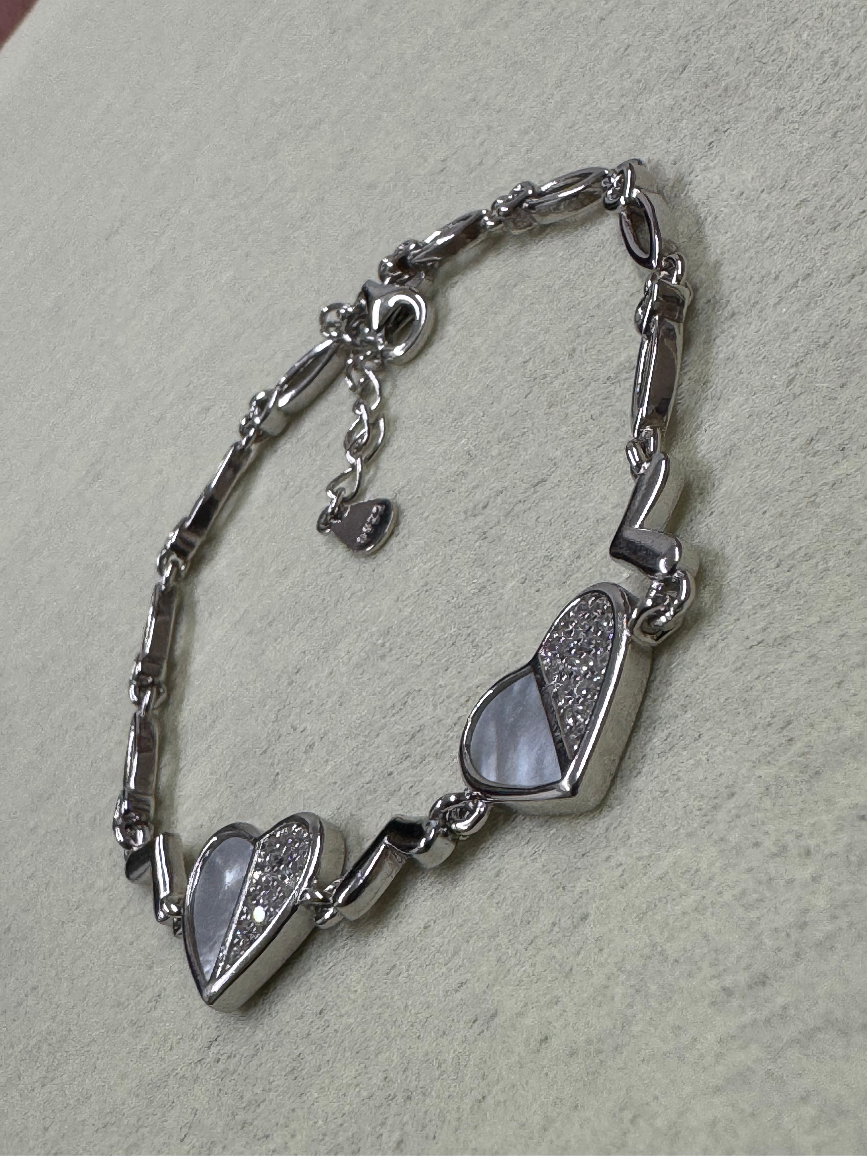 Sterling silver bracelet with two heart stations set with cubic zirconia, laid on woven beige fabric.