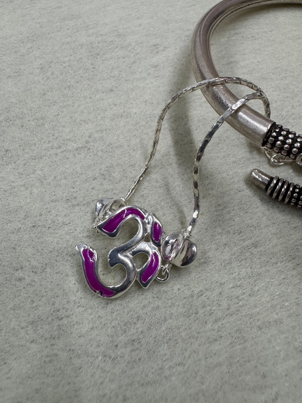 The magenta enamel Om station photographed on grey felt beside an oxidised silver bangle.