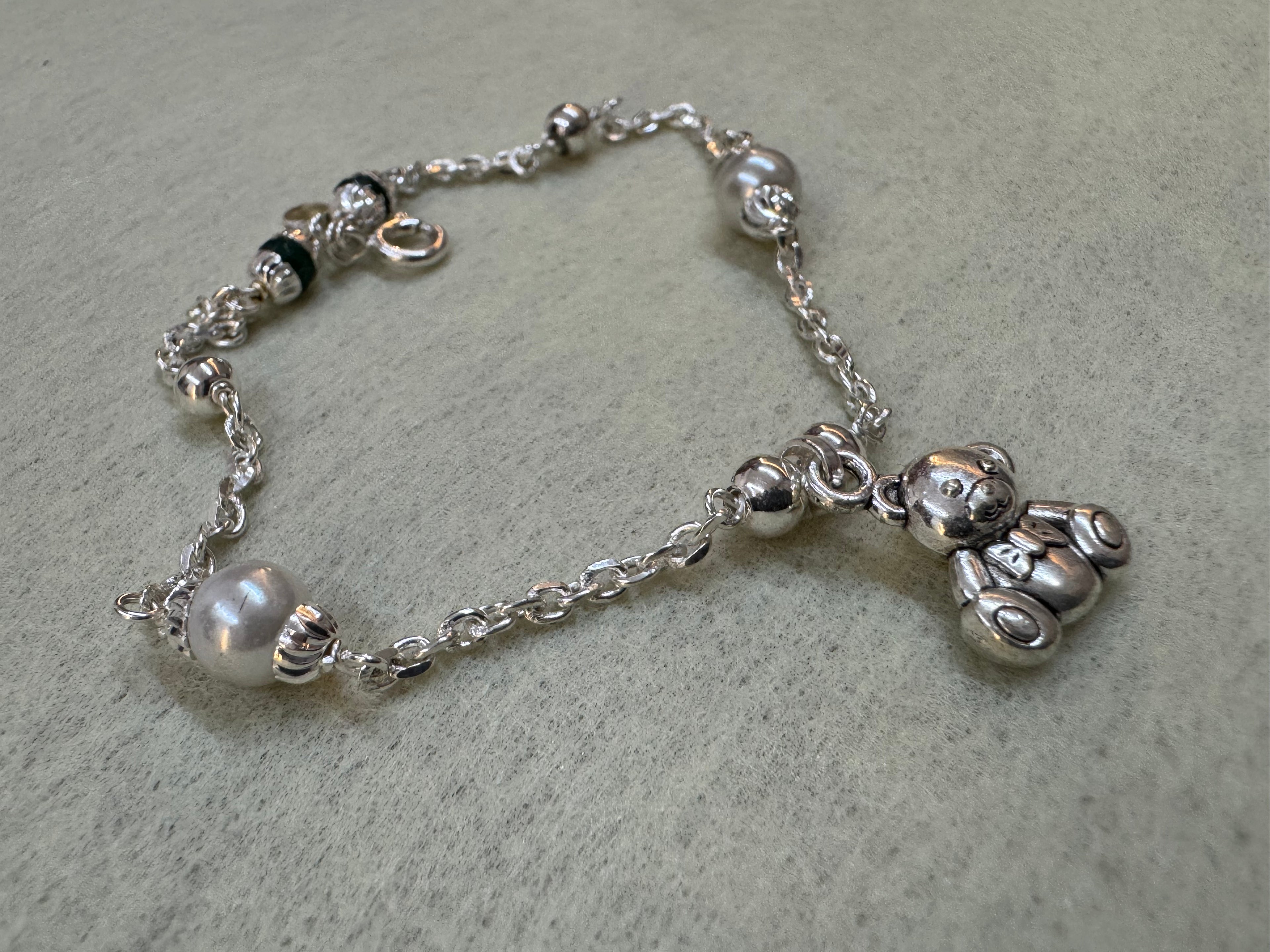 Sterling silver charm bracelet laid in a curve on grey, showing the bead stations, the teddy bear charm and the spring ring clasp.