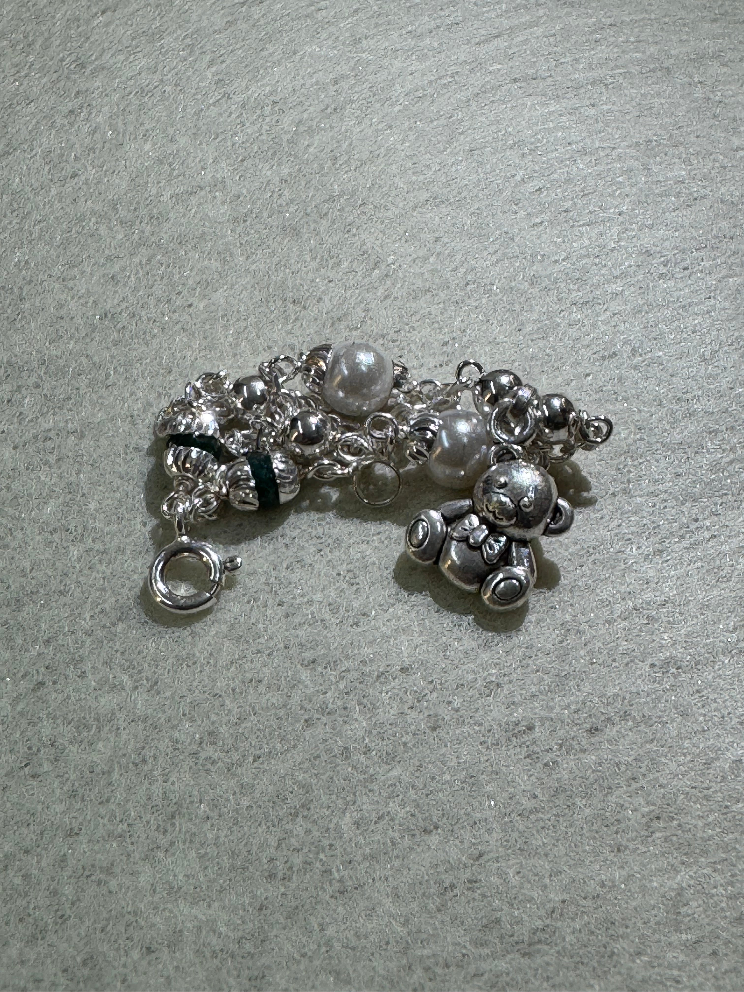 Sterling silver charm bracelet gathered into a loose coil on grey, with the teddy bear charm at the top and the spring ring below.