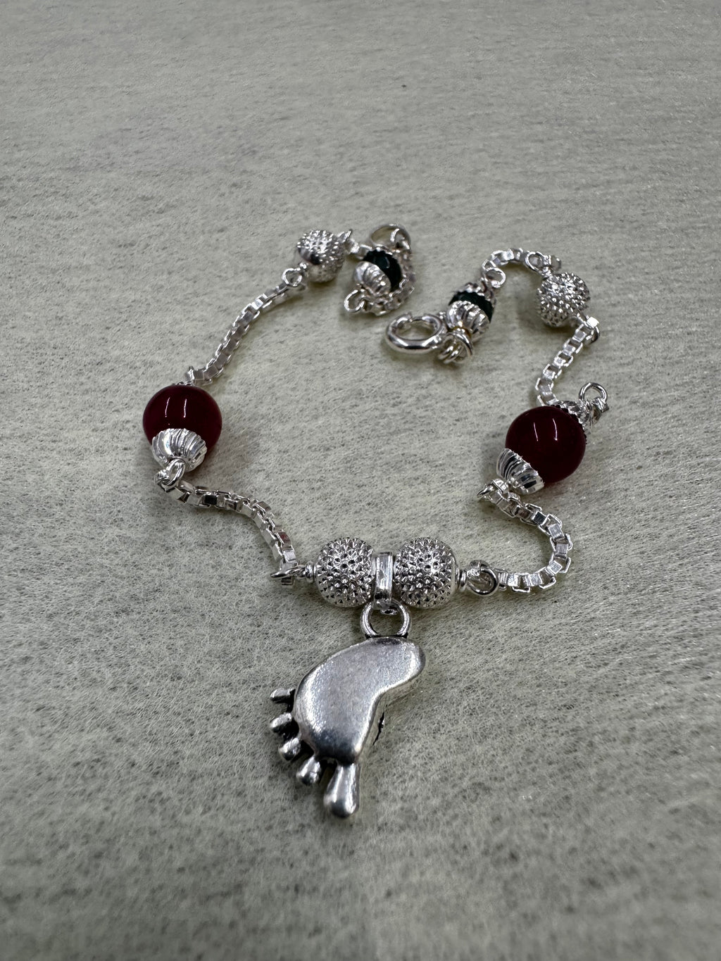 Sterling silver box chain bracelet with a cast baby foot charm, granulated silver balls and dark red capped beads.