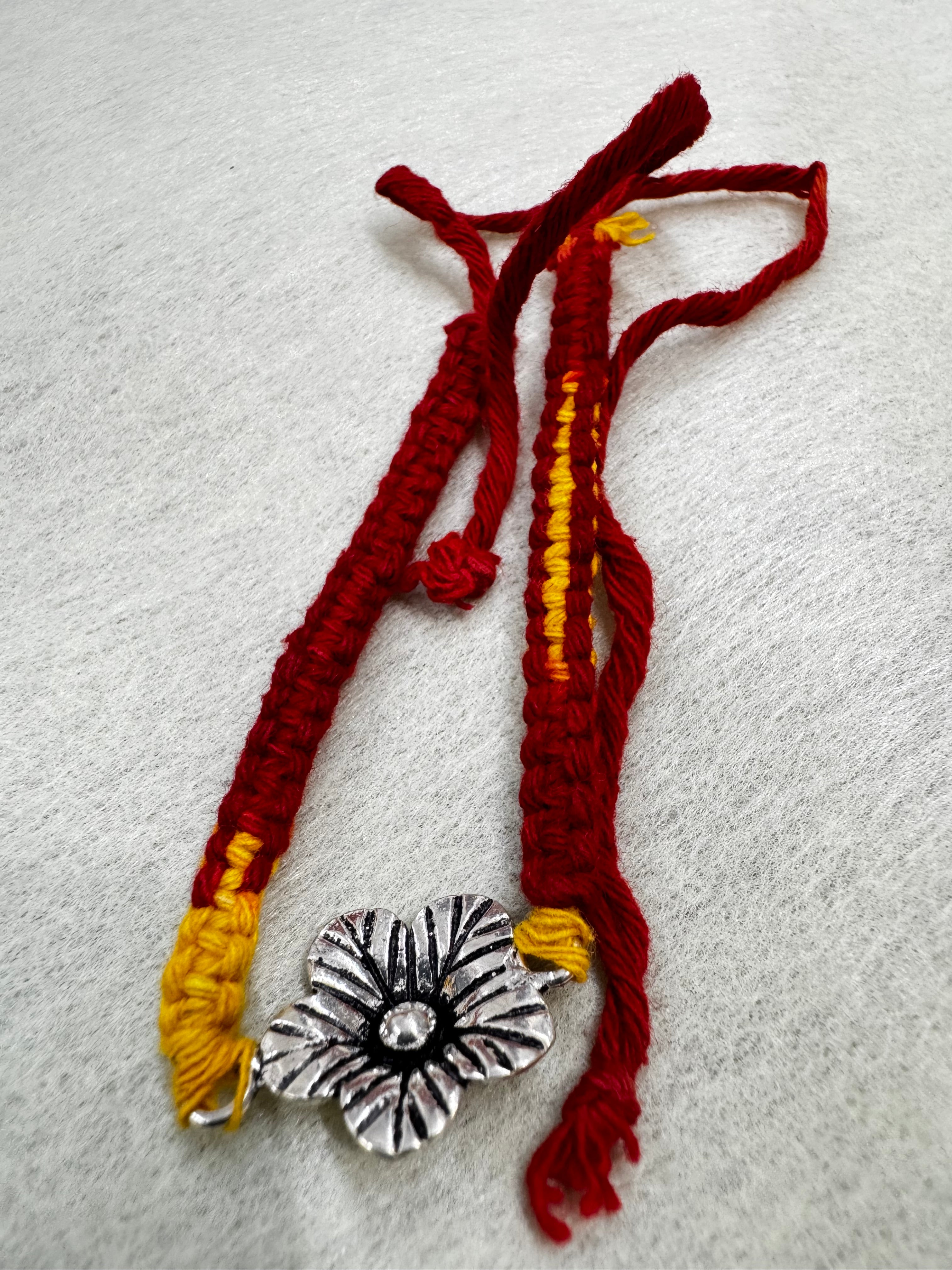 The full thread bracelet laid out straight, showing the sterling silver flower, the yellow stripe through the red band and the long ties.