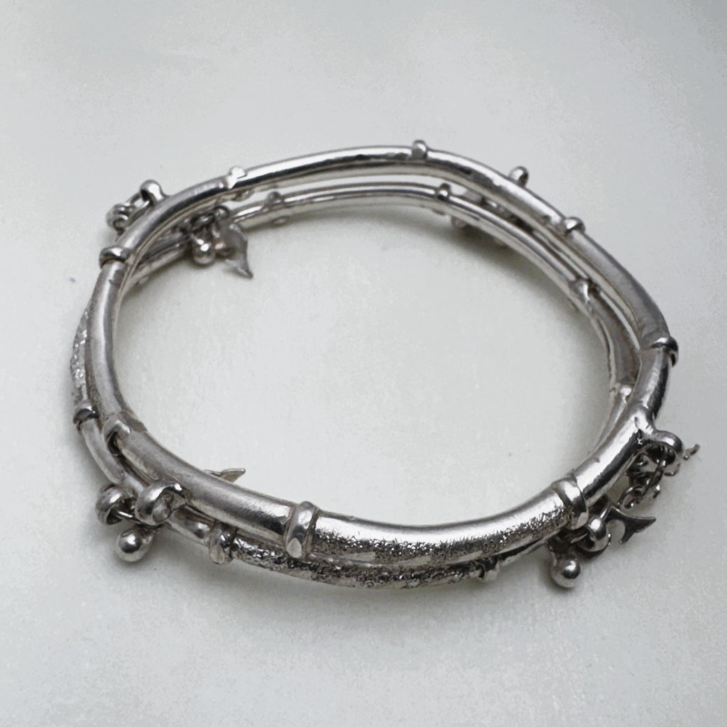 Sterling silver twin-tube kada with diamond-cut sections and small leaf and bead charms, seen at an angle.