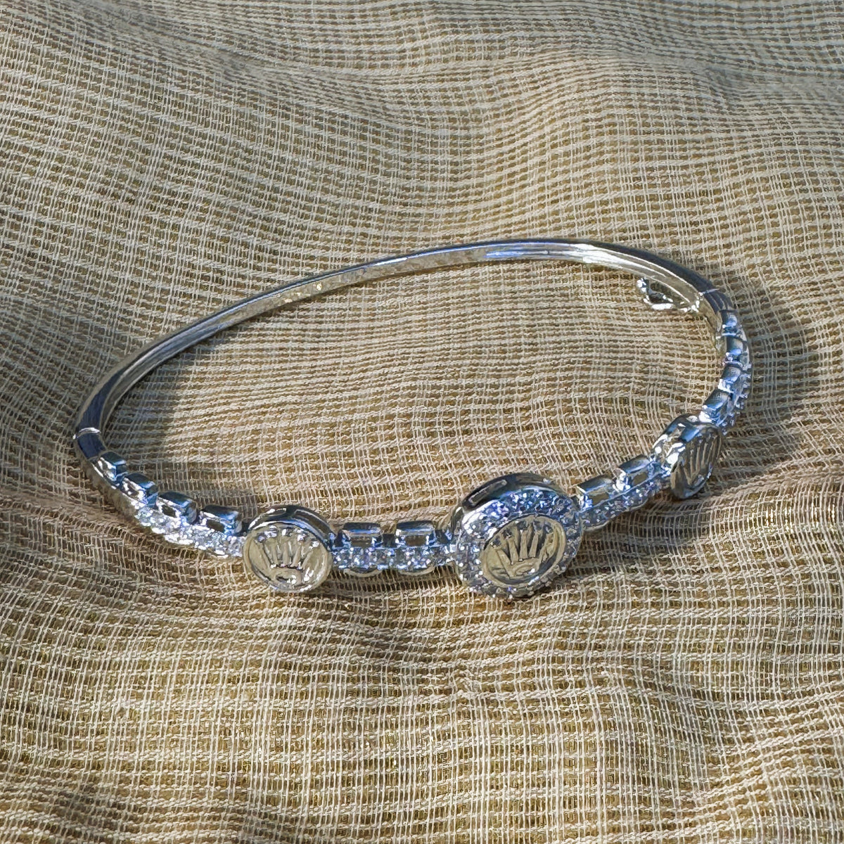 Sterling silver hinged bangle bracelet with three crown medallions and American Diamond pave links, on cream fabric.