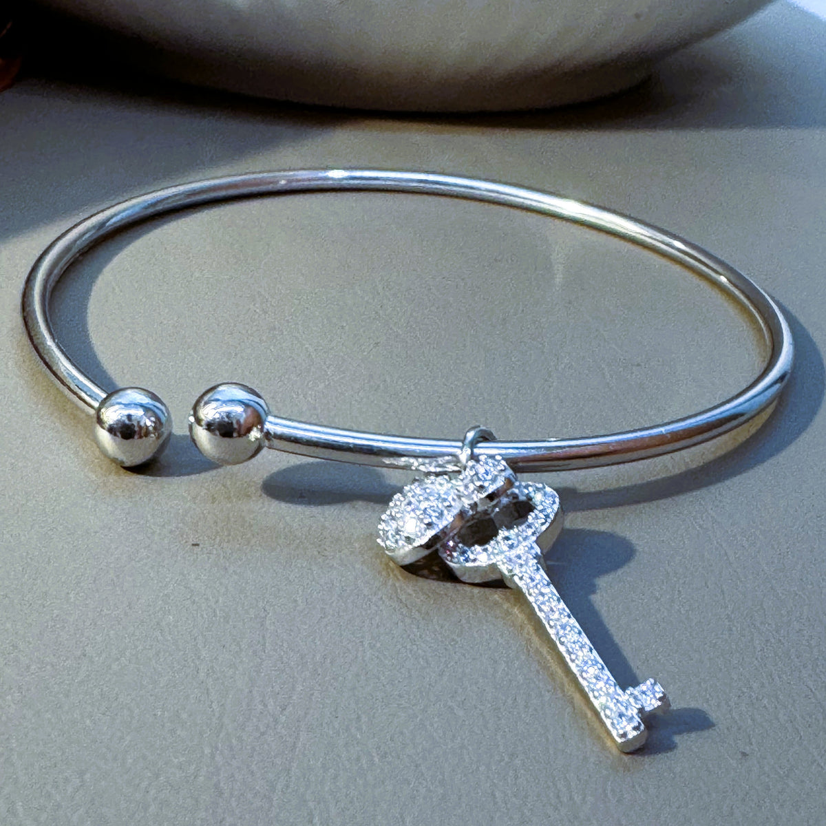 Sterling silver open cuff bracelet with ball ends, a pave key charm and a cartoon character charm, on a grey surface.