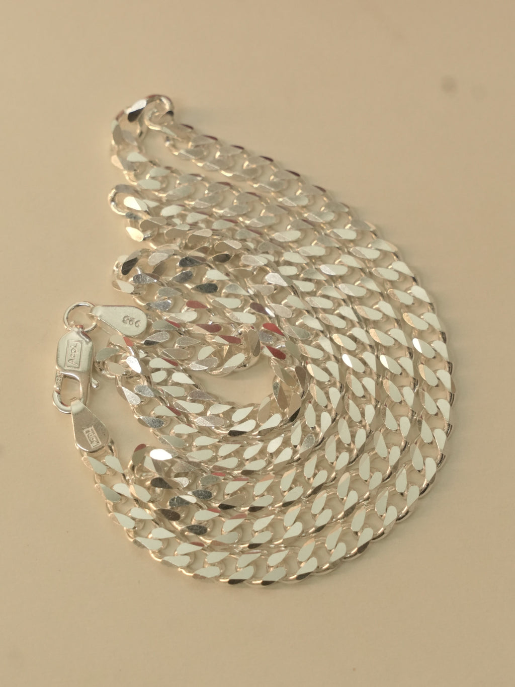 Sterling silver flat curb chain coiled on a pale surface, with the lobster clasp at the edge.