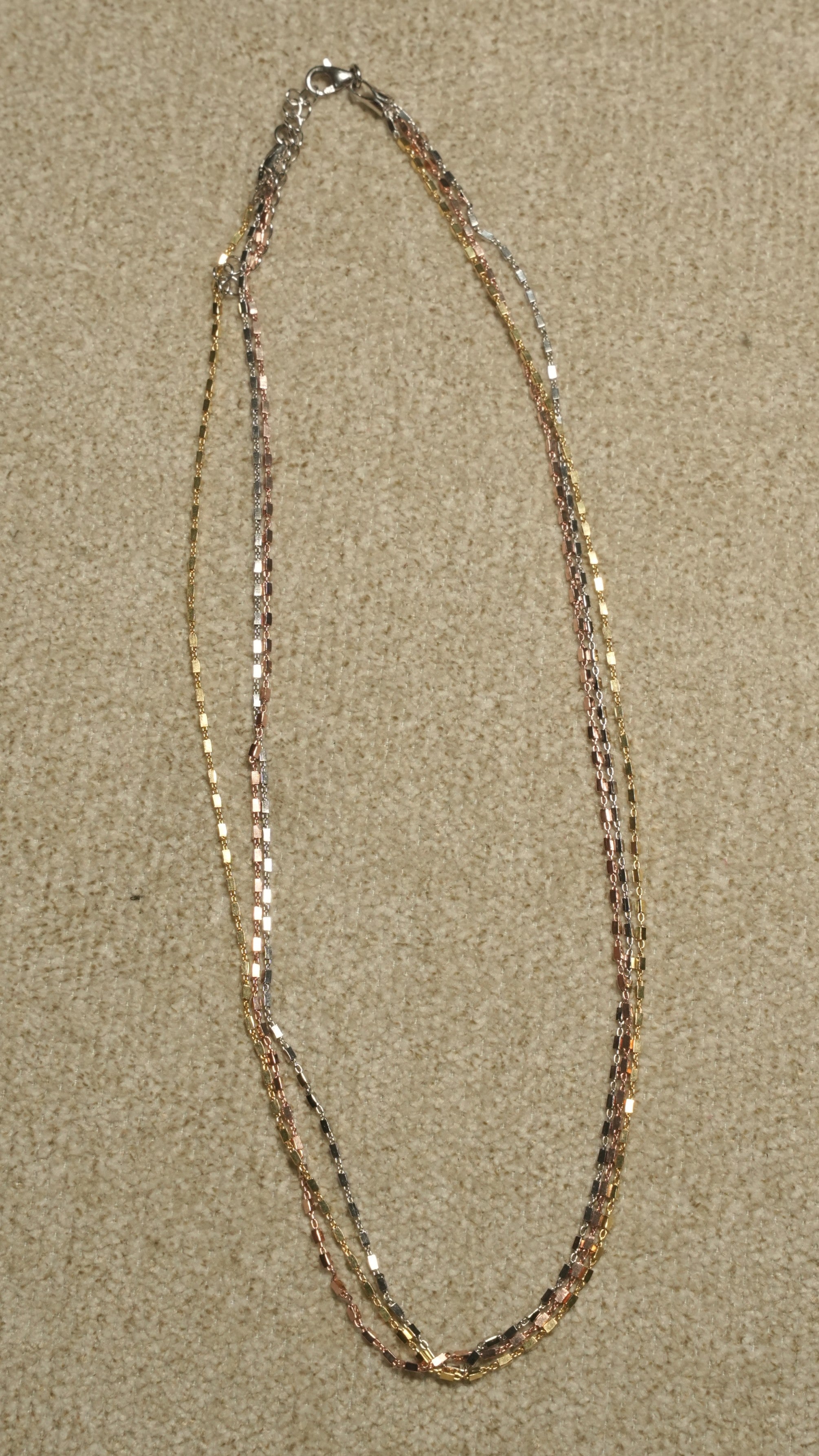 Triple-strand chain with silver, gold-tone and rose gold-tone faceted bar links, laid in an open oval on a beige textile backdrop.