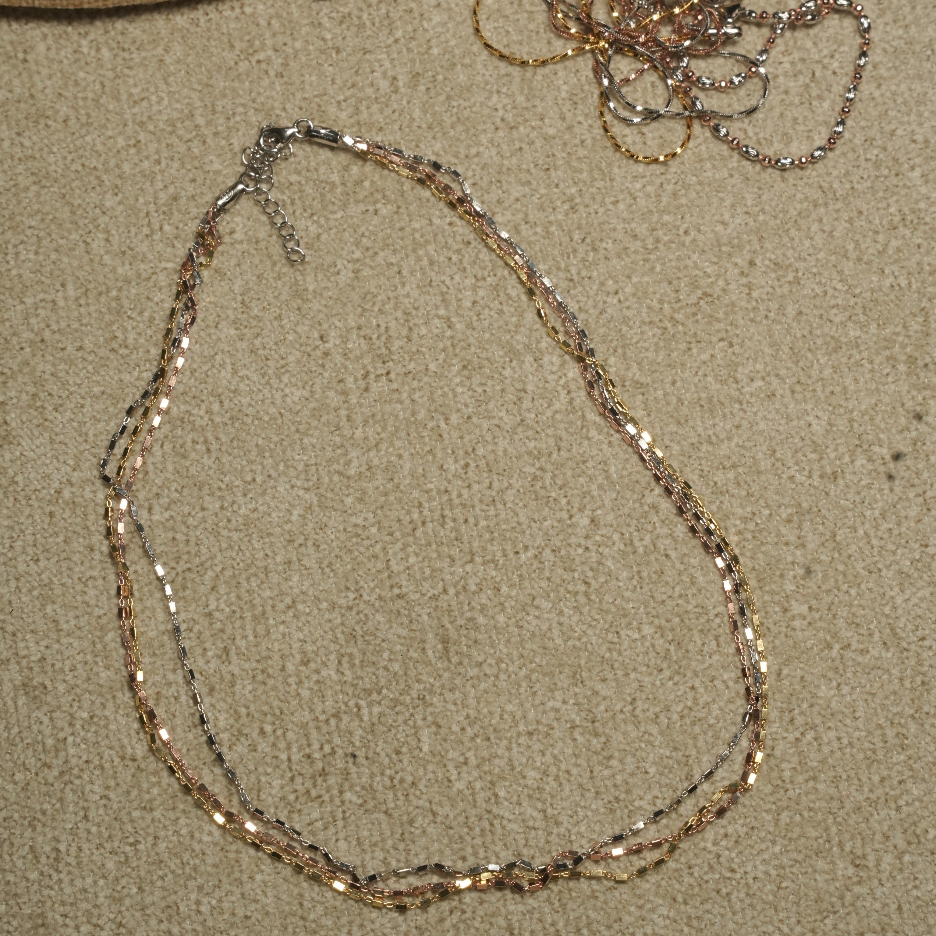 Tri-tone triple-strand sterling silver chain arranged in a loose loop beside a woven jute pouch, with the lobster clasp and extender chain visible.