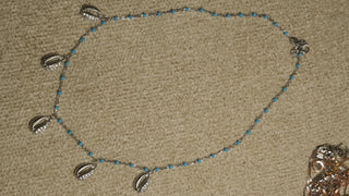 Sterling silver chain with turquoise-blue enamel beads and six cowrie shell drops set with cubic zirconia, laid flat on a beige woven backdrop.