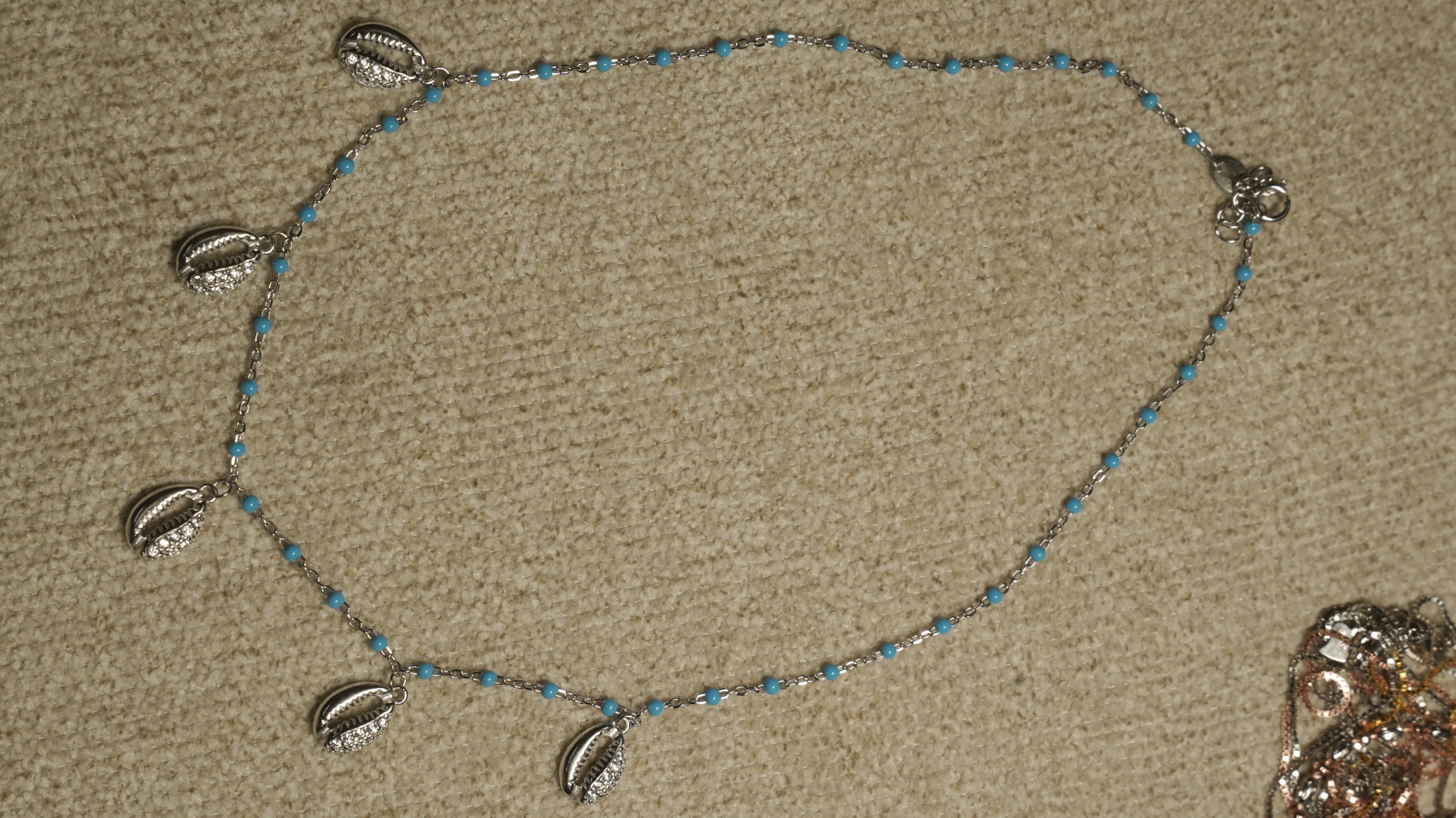 Sterling silver chain with turquoise-blue enamel beads and six cowrie shell drops set with cubic zirconia, laid flat on a beige woven backdrop.